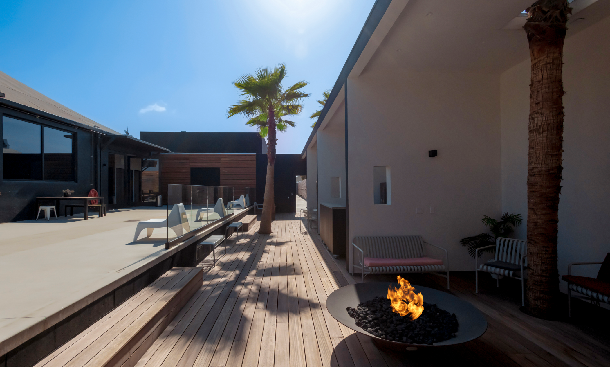 A modern style wooden deck with a fire pit, check chairs and palm trees.